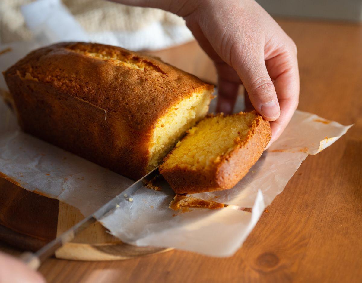 A hand slicing a fresh, golden pound cake in half on a wooden cutting board, revealing the moist interior
